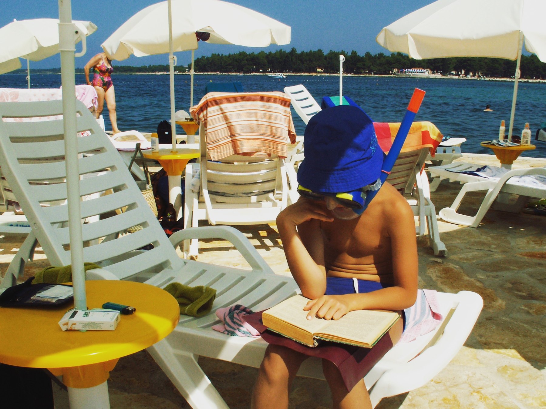 A person reading a book on a bright beach beside the sea.